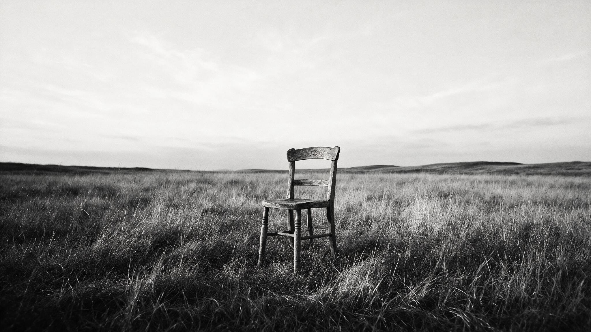 כיסא עץ ישן עומד לבדו בשדה עשב פתוח תחת שמיים בהירים. להיות עם מה שיש, בלי למהר לשנות